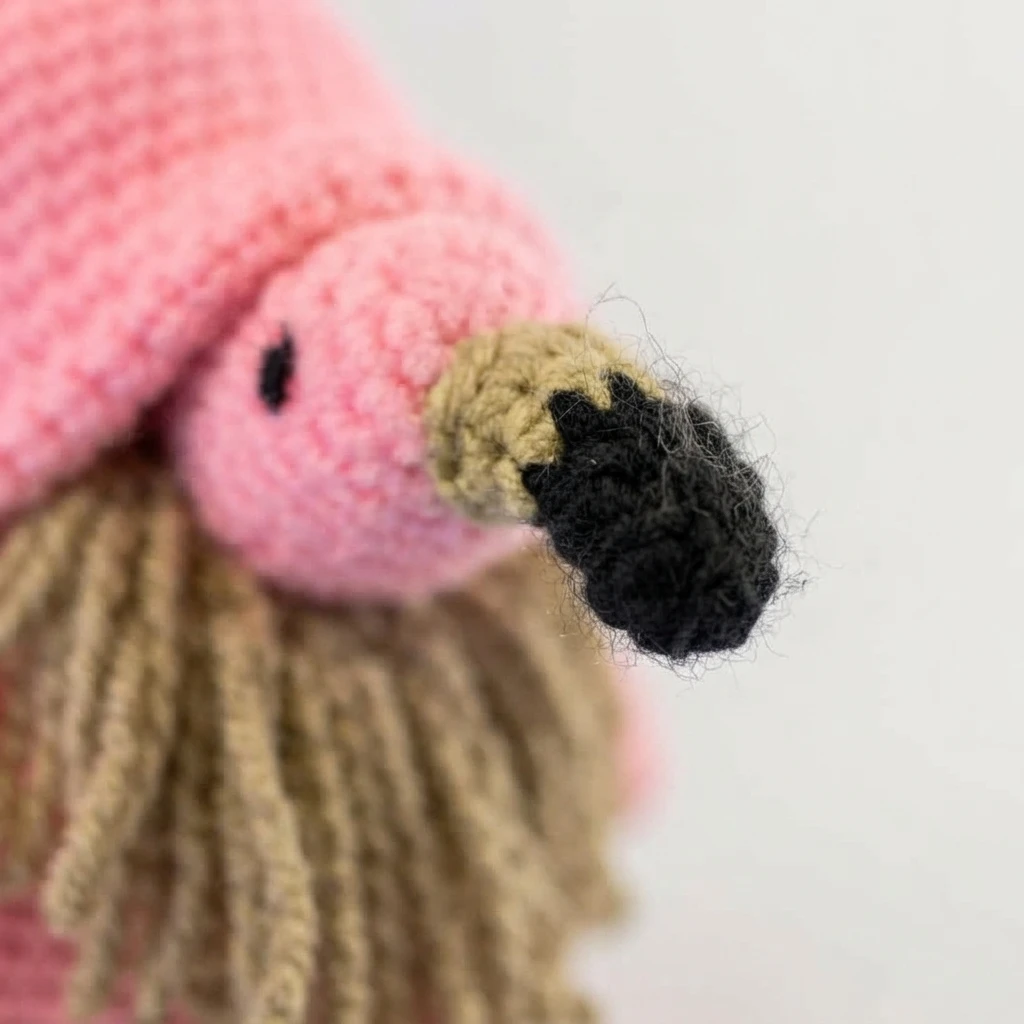 Close-up of a crocheted pink flamingo gnome head with a black stitched eye and a beak worked in buff and black yarn, framed above strands of tan yarn beard and a pink hat.