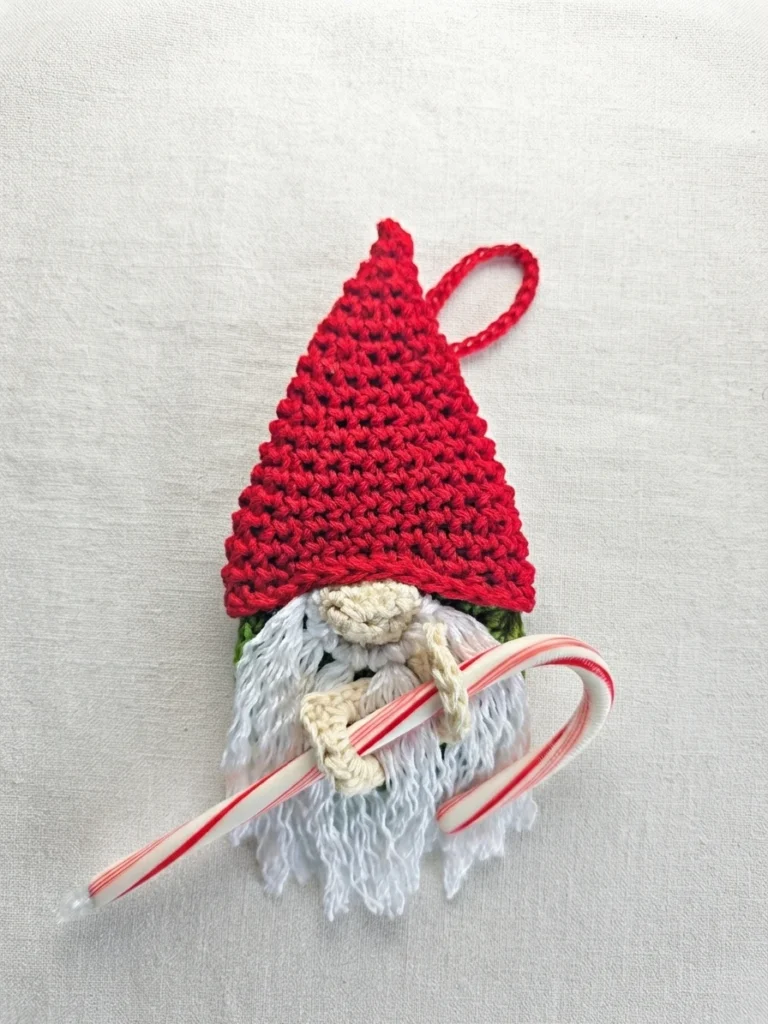 A crocheted gnome ornament in a red hat holds a red-and-white candy cane, with a hanging loop above the hat.