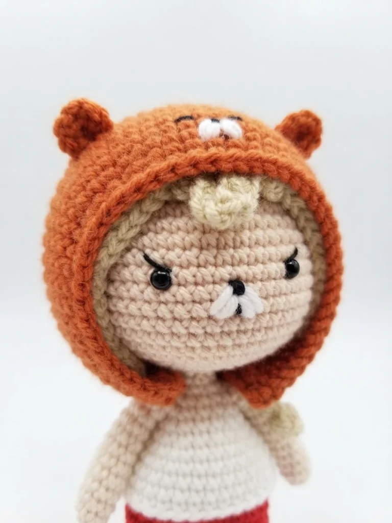 Close-up of a crochet amigurumi doll wearing an orange hamster hoodie with small ears, blonde hair, black eyes and a white top on a white background