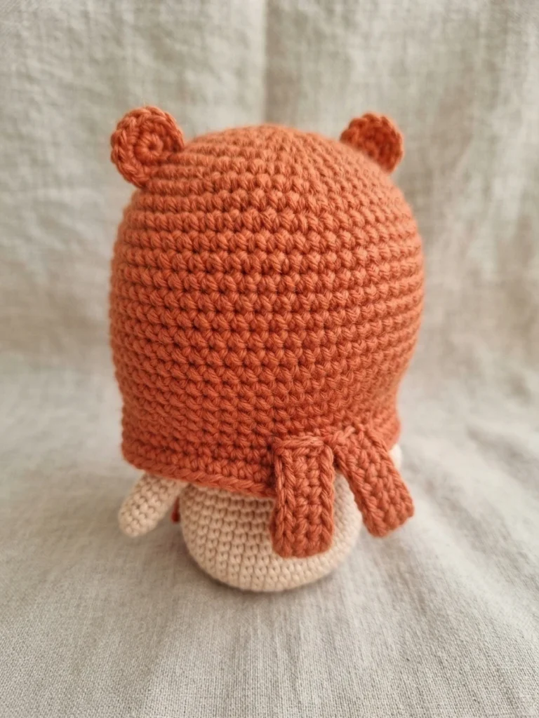 Back view of a crocheted doll wearing an orange hamster hoodie with small round ears and two hanging straps