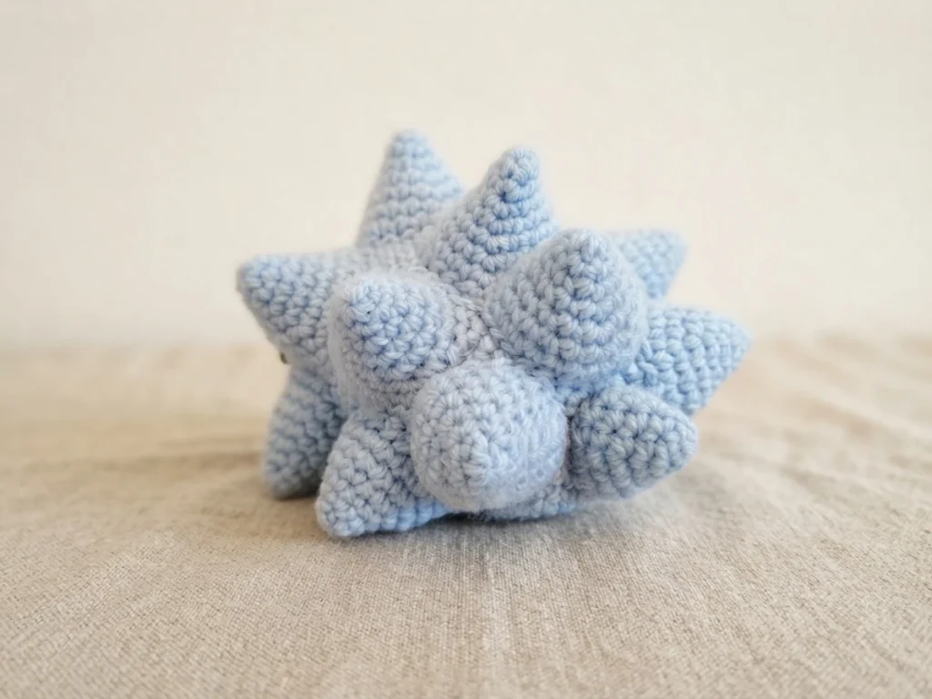 Rear view of the crocheted light blue spiky shell of the Snom amigurumi resting on a beige fabric surface.