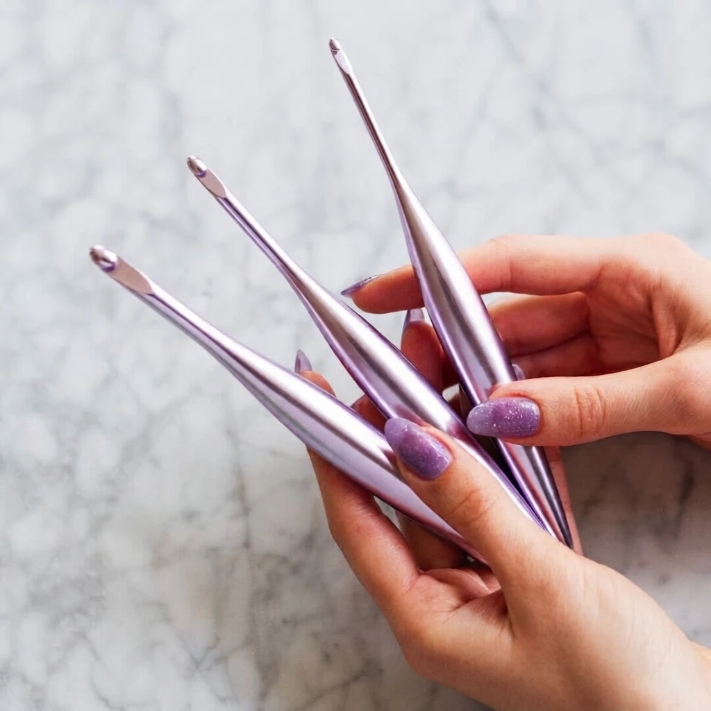 Hands holding three metallic lilac crochet hooks against a pale background.
