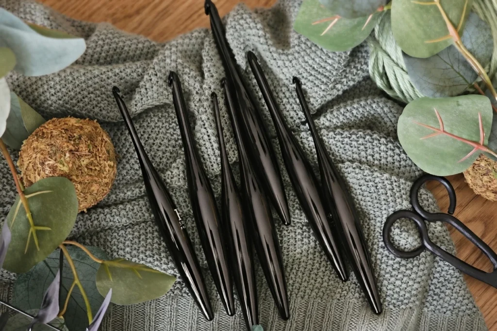 Black crochet hooks arranged on gray crocheted fabric beside scissors and decorative leaves.