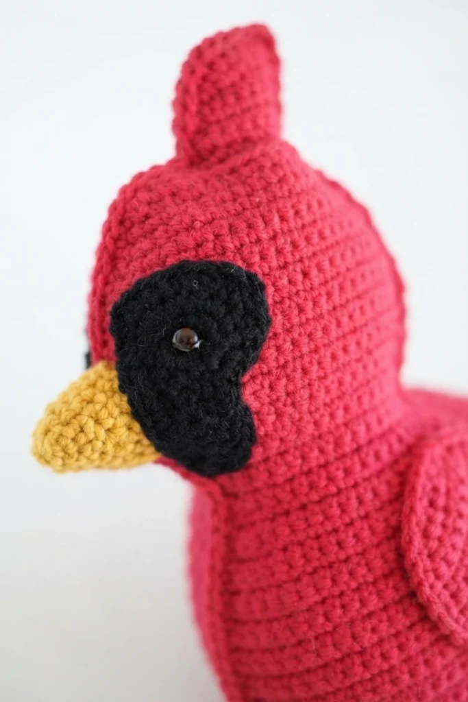 Red crocheted cardinal pillow with black face patch, yellow beak and crest, shown in close-up