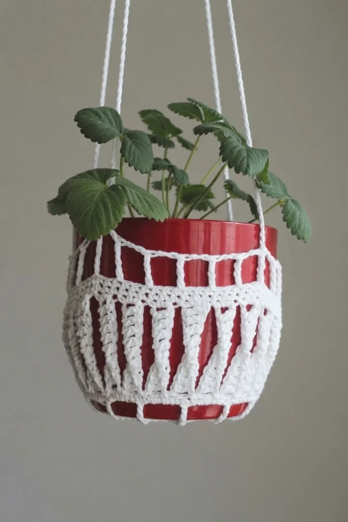 A white openwork crochet cover hangs around a red plant pot with leafy green foliage.