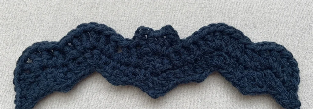 Dark navy crochet bat wing strip in progress with a wavy scalloped edge and a raised section in the middle, laid flat on a light textured fabric surface.