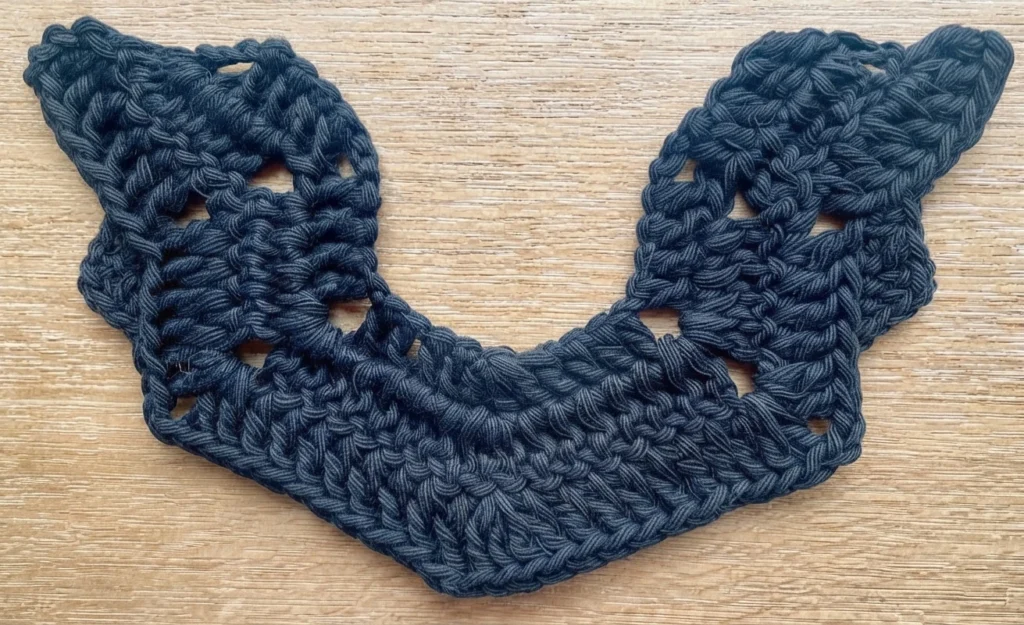 Dark blue crocheted bat wing shape with wavy edges, open chain spaces along the wings and a V shaped dip in the middle, lying flat on a light wood grain surface.