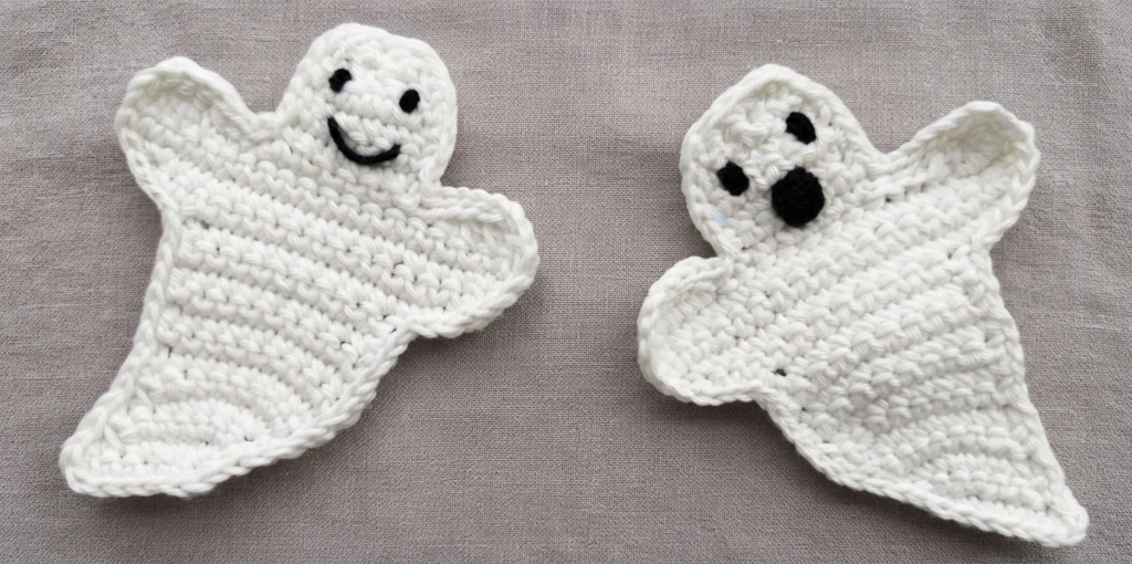 Two white crochet ghost appliqués with black embroidered faces, one smiling with two round eyes and a curved mouth, the other with a round eye and an open oval mouth, placed side by side on a plain grey fabric background.