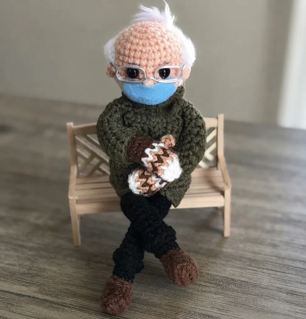 Crocheted doll of an elderly person with white hair, glasses and a blue face mask, sitting on a miniature wooden bench with hands resting on a brown and cream striped crocheted piece.