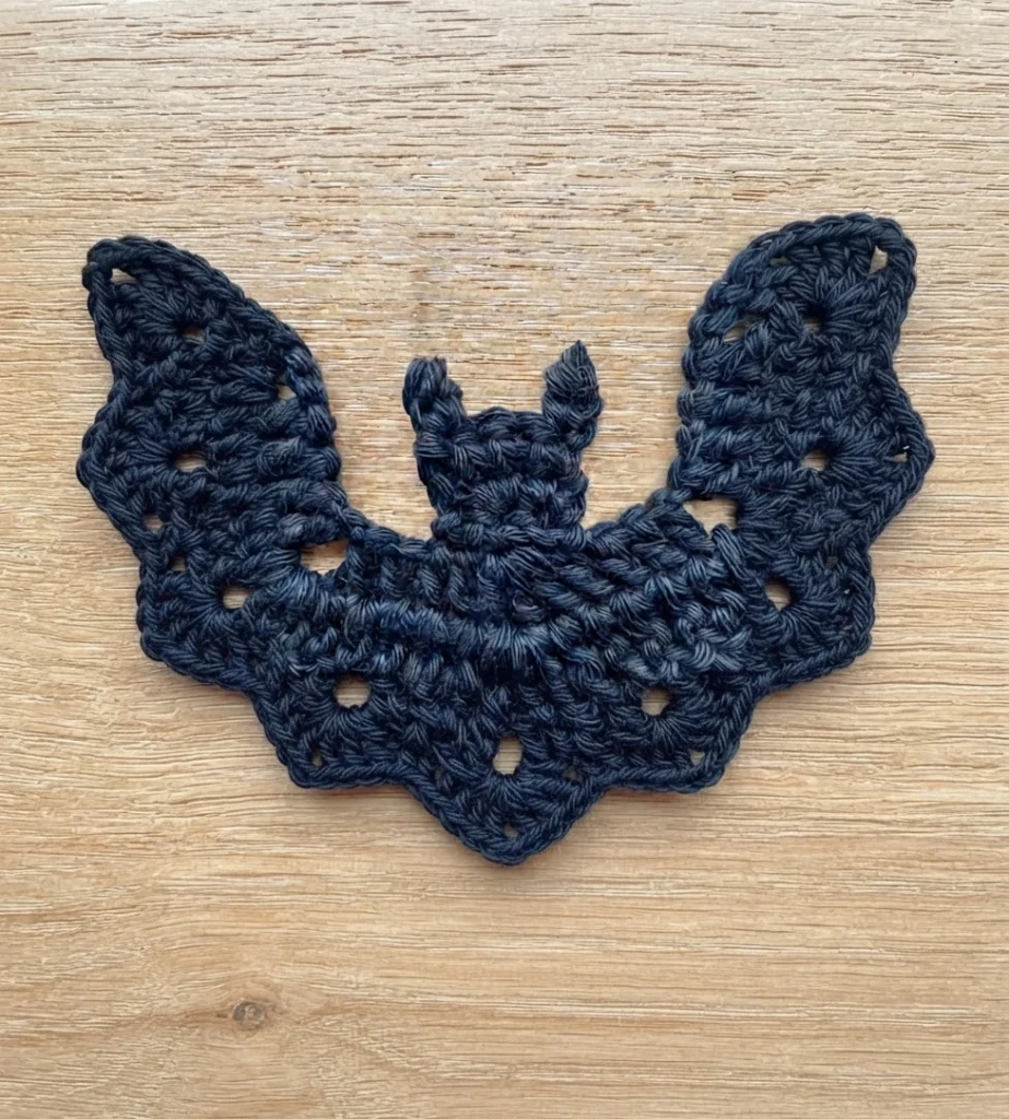 Dark navy crochet bat appliqué with two short pointed ears and wide scalloped wings, laid flat on a light wood surface.