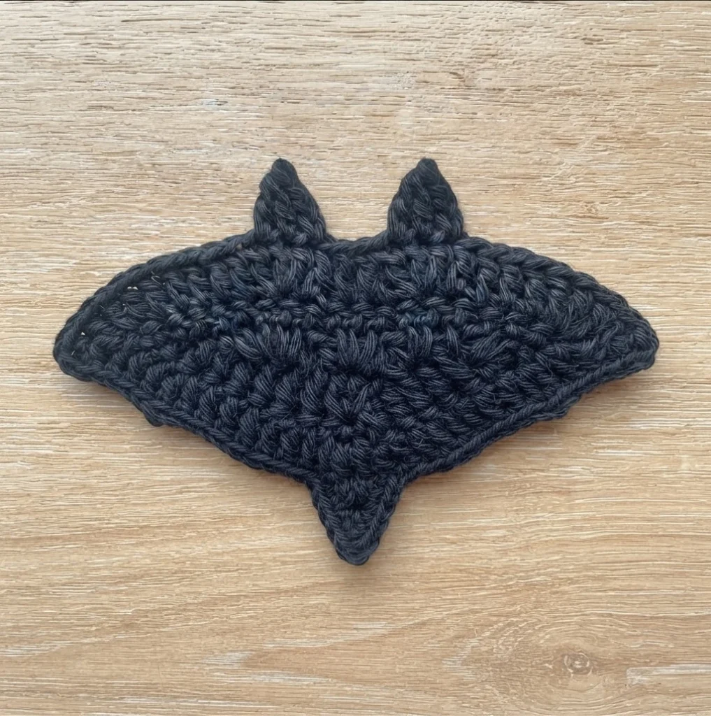 Dark charcoal crochet bat appliqué with two pointed ears and a pointed bottom tip, lying flat on a light wood surface, shown as a finished motif.