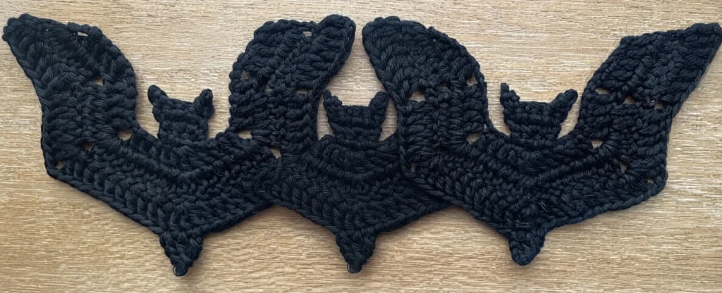 Three dark navy crocheted bat appliques joined edge to edge on a light wood grain surface, each with pointed ears, wavy openwork wings and a pointed lower tip