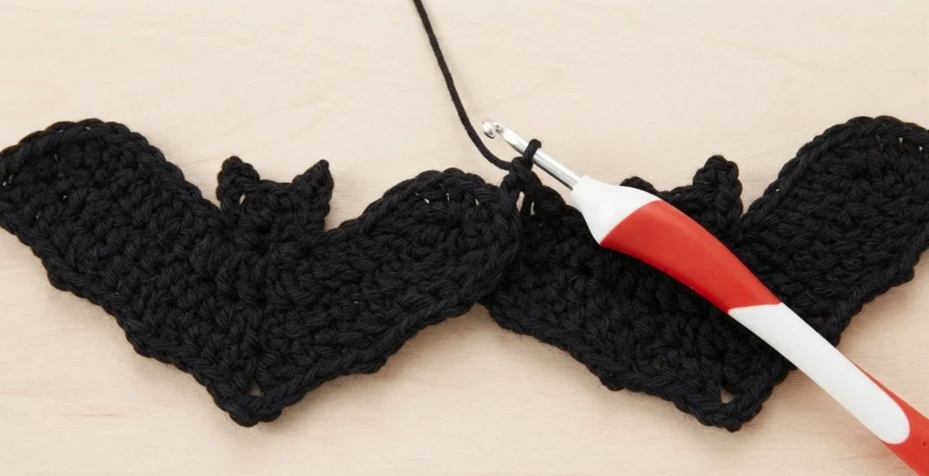 Two black crocheted bat shapes lying side by side on a pale wood surface, with a red and white handled crochet hook resting at the point where their top edges meet and a black yarn strand rising out of frame.