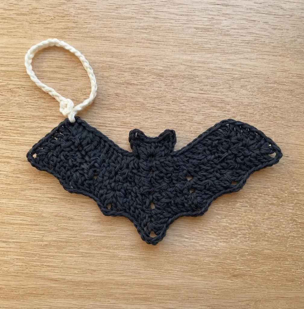 Black crochet bat with pointed ears and a cream chain hanging loop attached at the tip of one wing, laid flat on a light wood surface