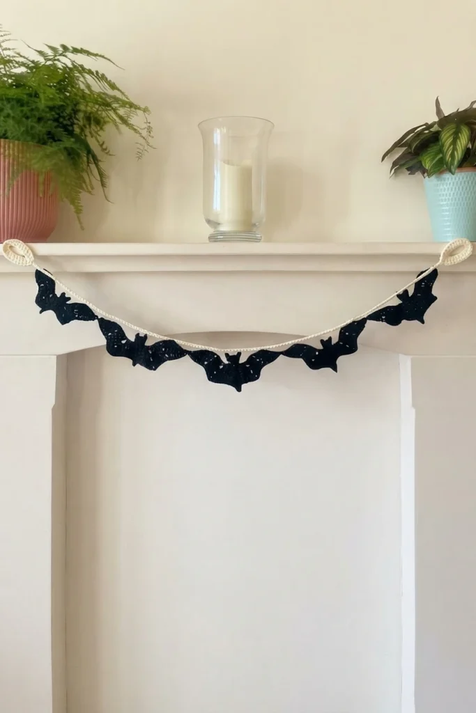 Black crochet bat garland hanging on a white mantelpiece, suspended from a cream crocheted cord with a small loop at each end, with potted plants and a glass candle holder on the shelf above.