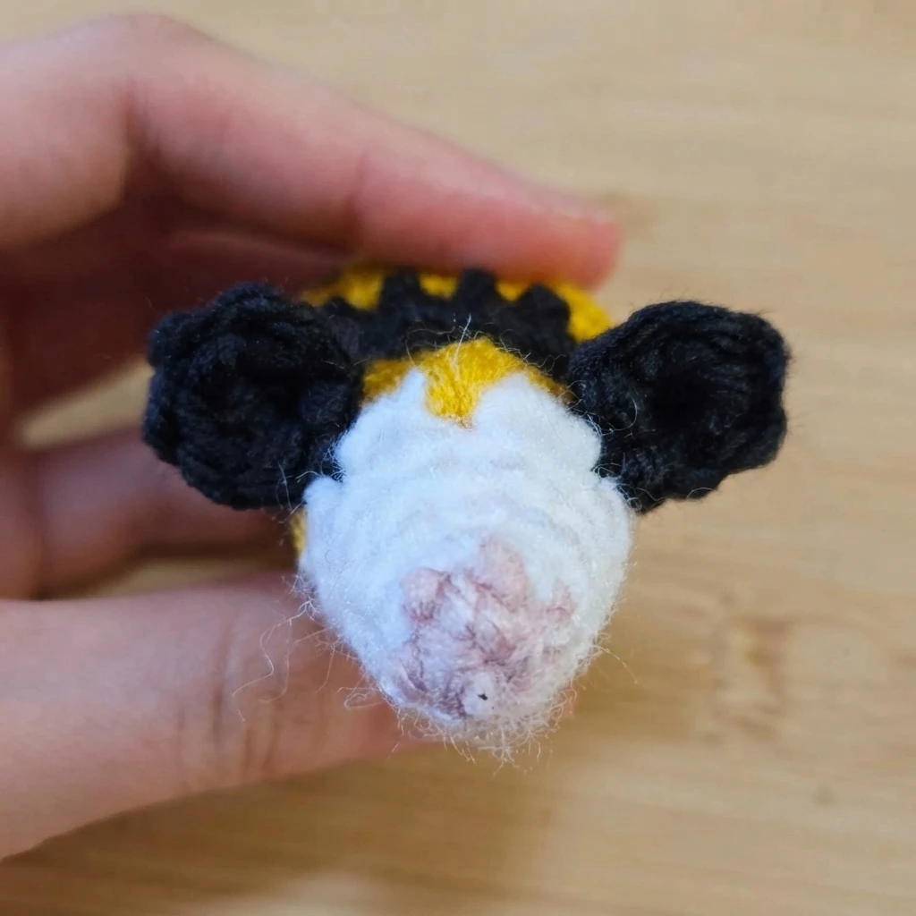 Hand holding a small crocheted bee head with black ears, a white face, a pale pink nose and a yellow and black striped body, on a wooden surface.