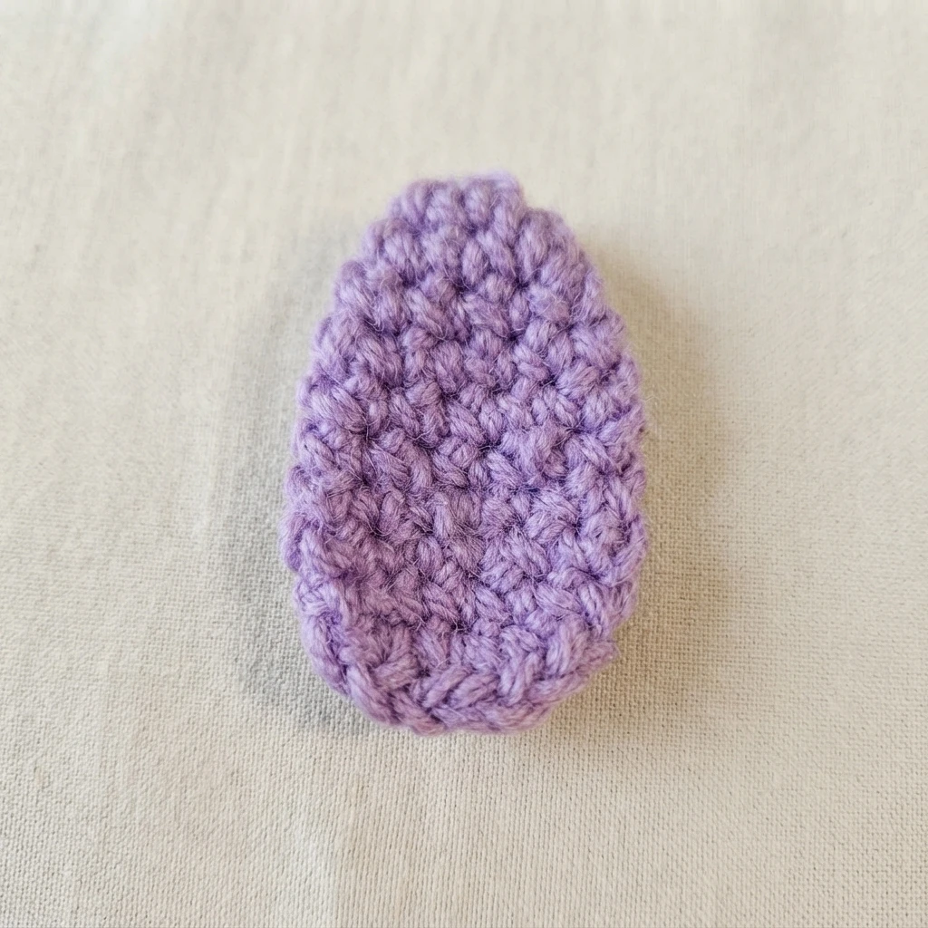 Single light purple crocheted petal piece lying flat on a plain beige fabric background