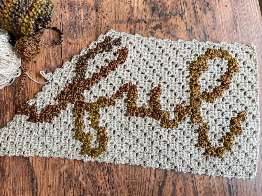 Gros plan d’un chemin de table au crochet crème avec des lettres brunes et ocre, accompagné de pelotes de laine sur une table en bois.