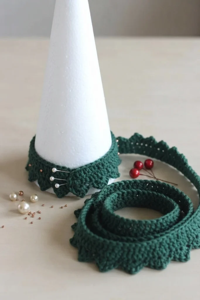 A white foam cone standing on a pale wood surface with a dark green crocheted ribbon wrapped around its base and pinned in place with round headed pins, next to a coiled crocheted ribbon with a scalloped edge, loose pearl and gold beads and