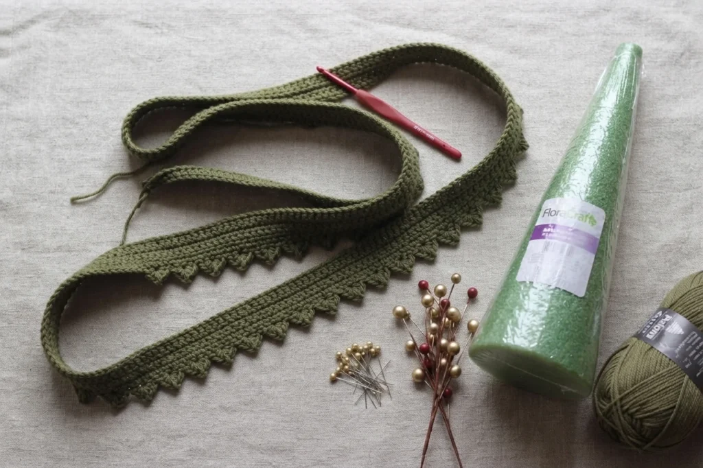 A long crocheted olive green ribbon with a scalloped picot edge coiled on a light fabric surface beside a red crochet hook, a green floral foam cone in plastic wrap, gold and red berry picks, a small pile of pearl headed pins and a ball of