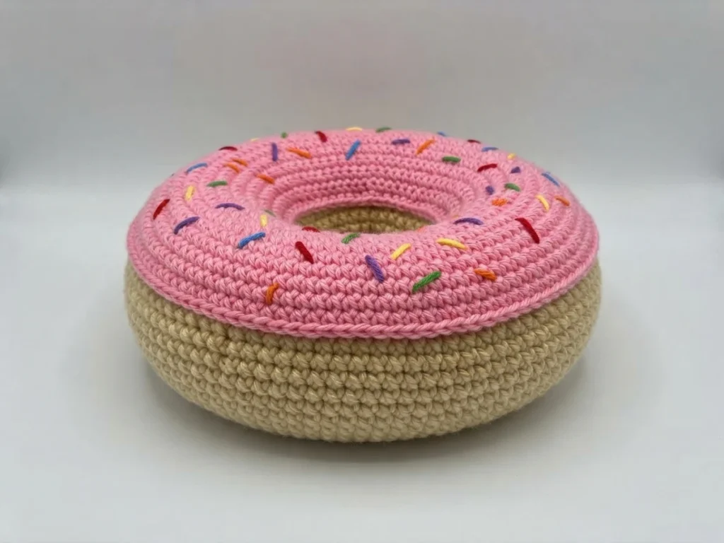Crocheted donut cushion with pink frosting and colorful sprinkles on a white background.