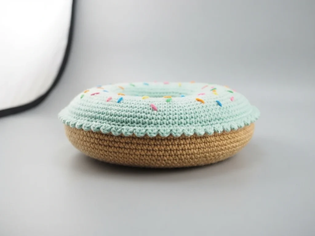 Crocheted donut cushion with pale green frosting, small colorful sprinkles and tan base on a plain background