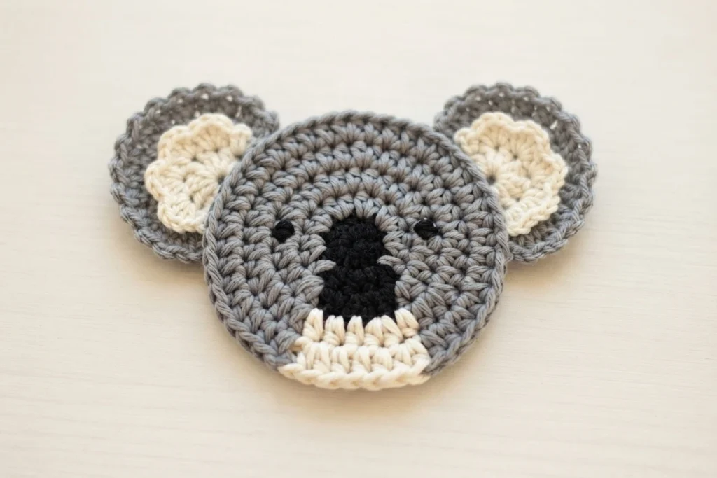 Grey crocheted koala coaster face with black nose, cream chin and cream inner ears