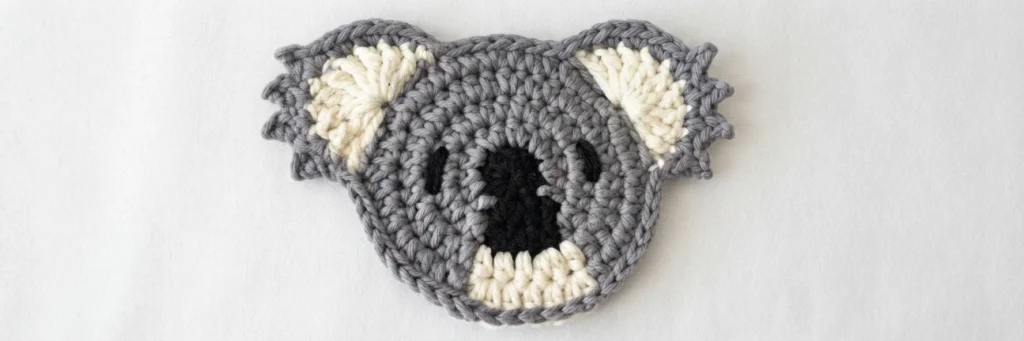Crocheted grey koala coaster with cream inner ears and chin, black nose and small embroidered eyes on a white background.
