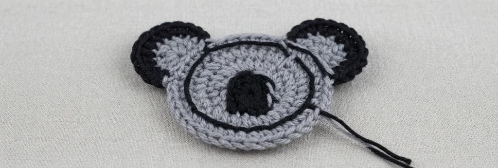 Partially crocheted grey koala coaster with black ears and nose, showing a strand of black yarn carried around the center on a light fabric surface