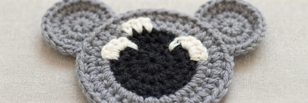 Close-up of a grey crochet koala coaster with a black oval nose and cream eye details resting on light beige fabric