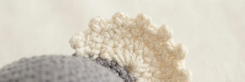 Close-up of a cream crocheted koala ear with small picot bumps along its edge, beside grey crochet fabric