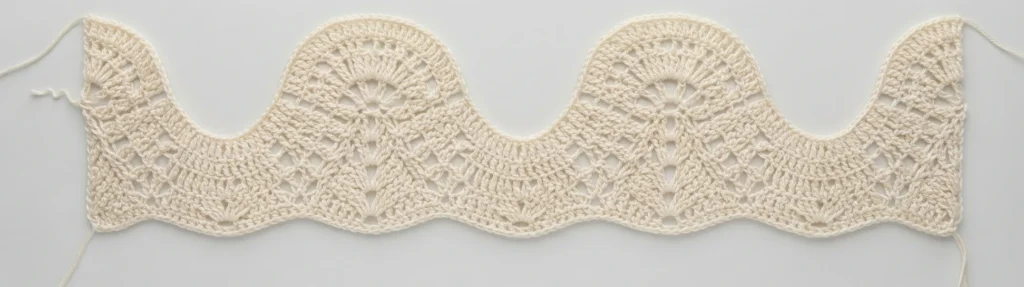 Cream-colored crochet swatch showing a repeating feather and fan wave motif worked in rows, with scalloped top and bottom edges and loose yarn ends at both sides, laid flat on a pale background.
