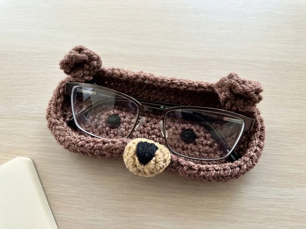 Brown crochet teddy bear eyeglass holder with a beige muzzle and black nose holding eyeglasses on a light wood surface
