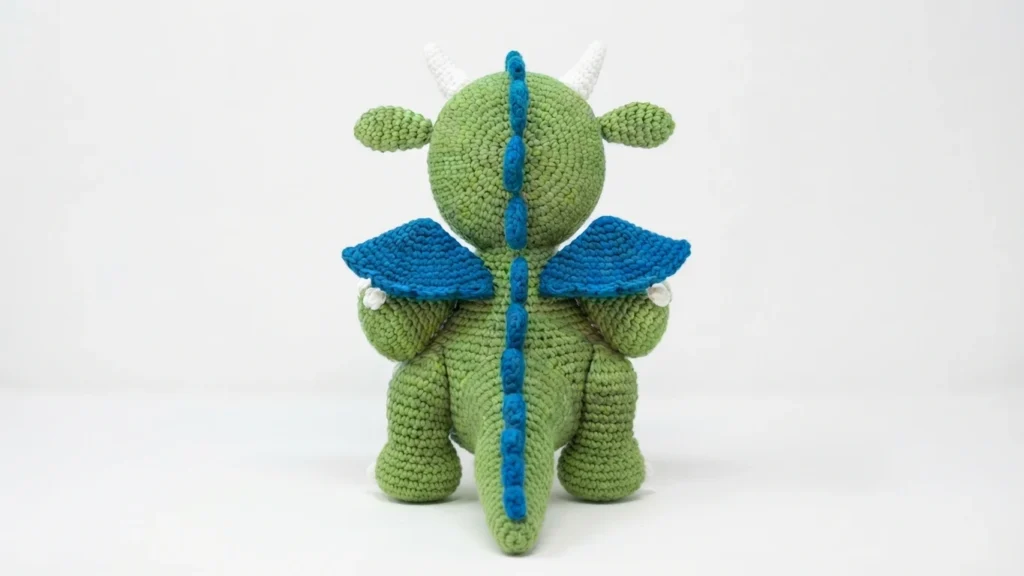 Back view of a green crocheted dragon with blue wings and a blue spiked ridge along its back and tail