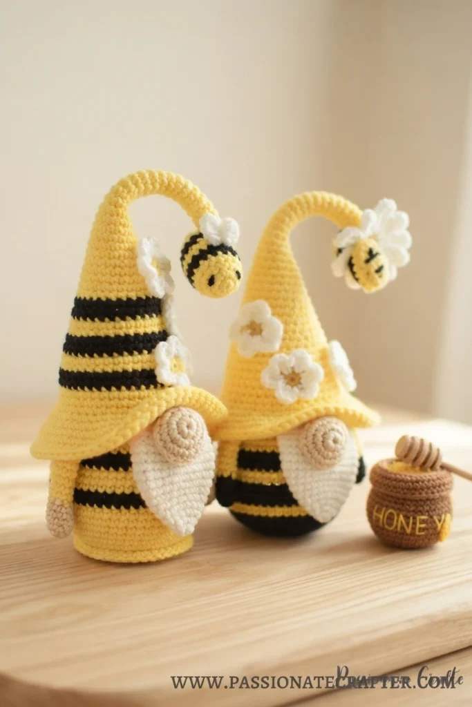 Two crocheted bumble bee gnomes with yellow hats, black stripes, white beards and flower appliques, next to a small honey pot