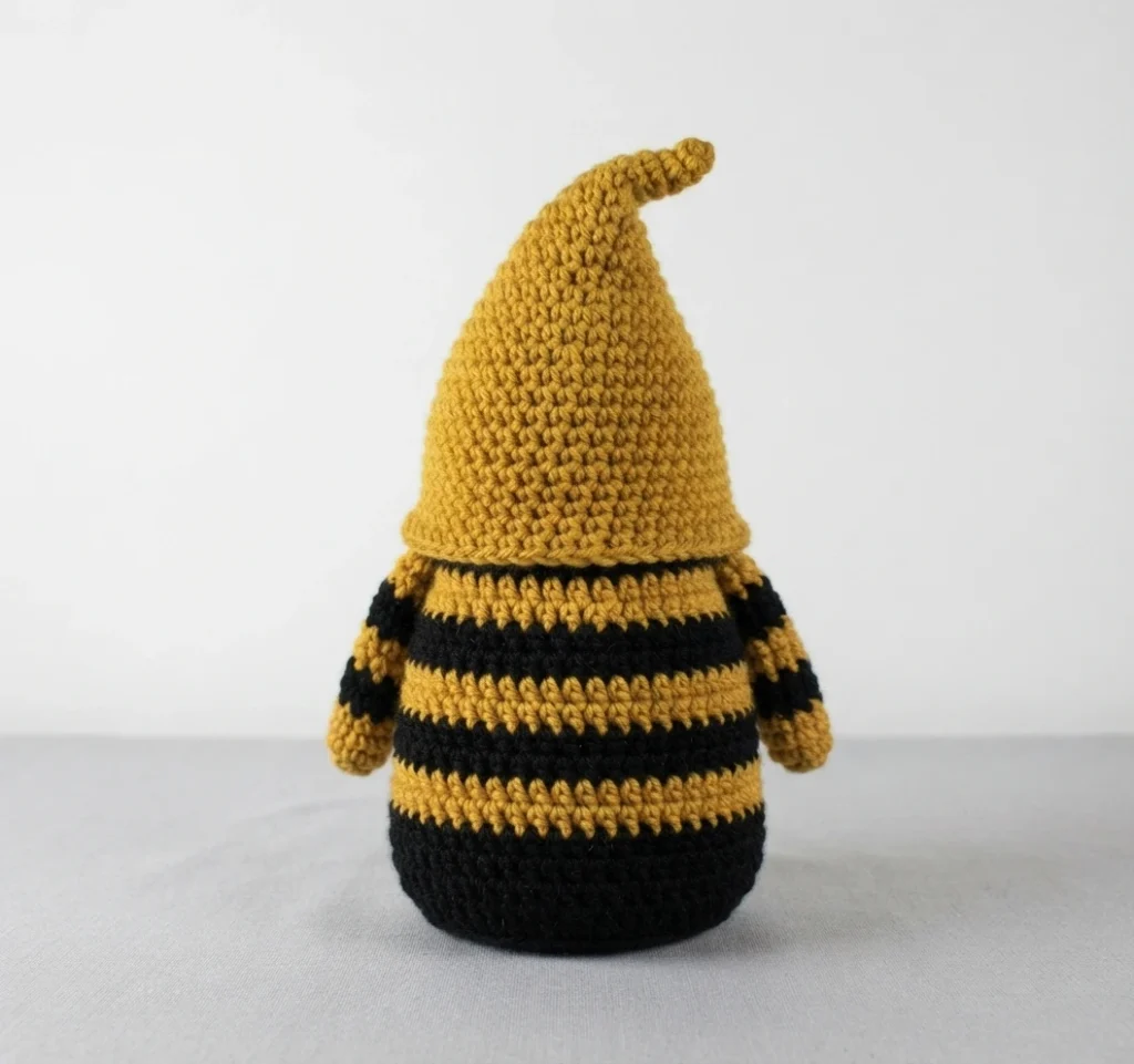 Back view of a crocheted bumble bee gnome with solid mustard yellow pointed hat and black and mustard striped body