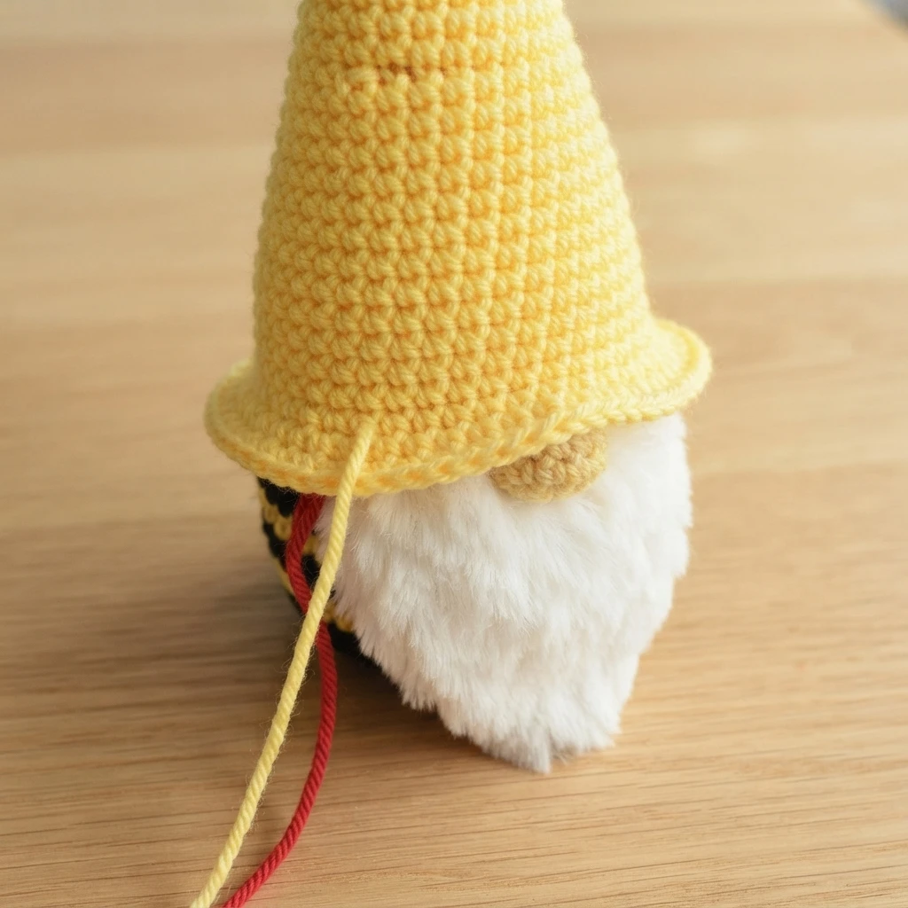 Finished crochet gnome with a tall yellow pointed hat and flared brim, white fluffy beard, yellow and red yarn tails trailing, on a wooden surface.