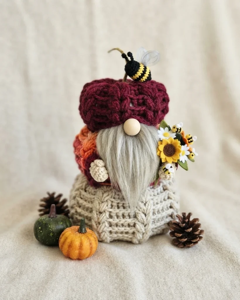 Crocheted gnome with burgundy, orange and cream textured body, long grey beard, wooden nose, sunflower and daisy bouquet with small bees, pinecones and mini pumpkins on beige fabric