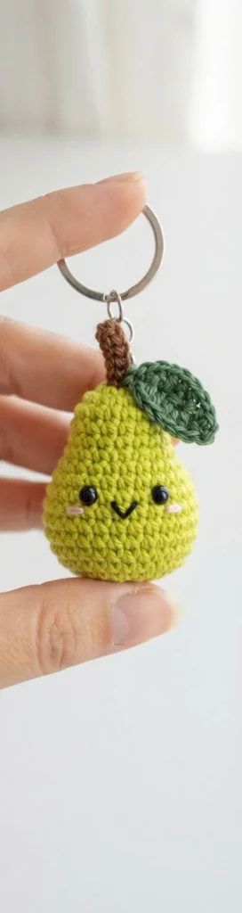 Small green crochet pear keychain with embroidered face, brown stem, dark green leaf and metal ring, held between two fingers