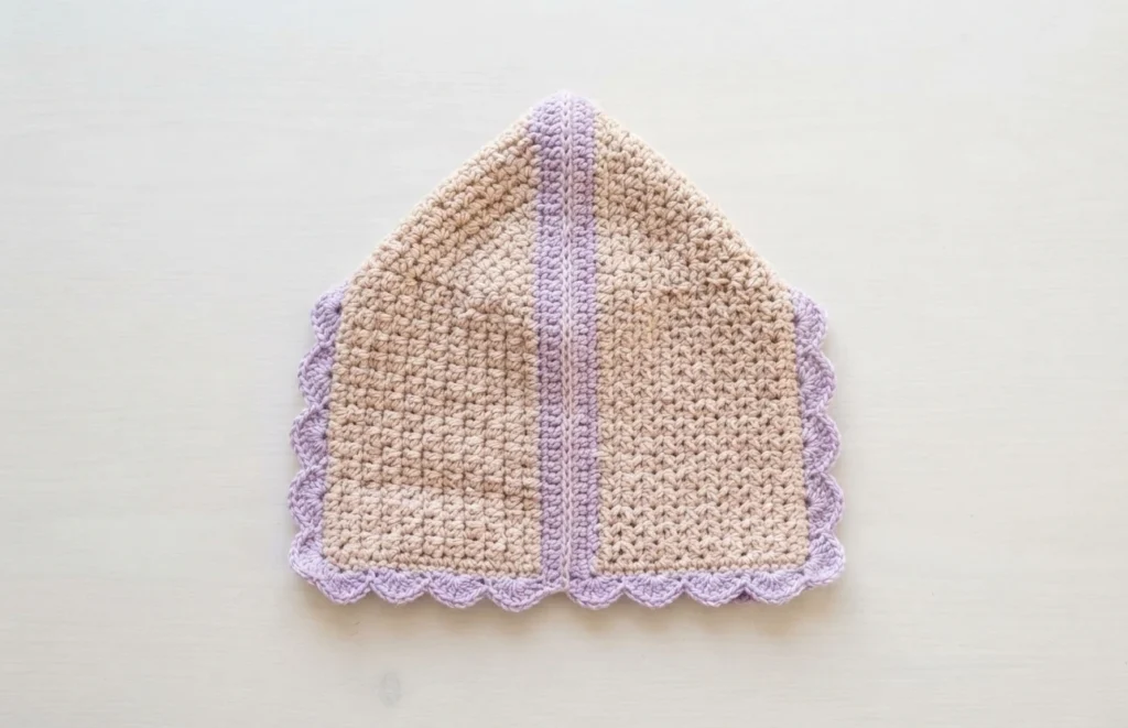 Seamed crochet pixie bonnet body laid flat in beige with lavender scalloped edging