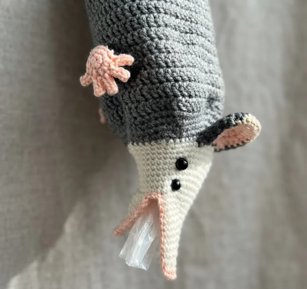 Crocheted gray opossum bag dispenser hanging with a white snout, pink paw raised and a plastic bag emerging from its open mouth, shown against a soft beige fabric background.