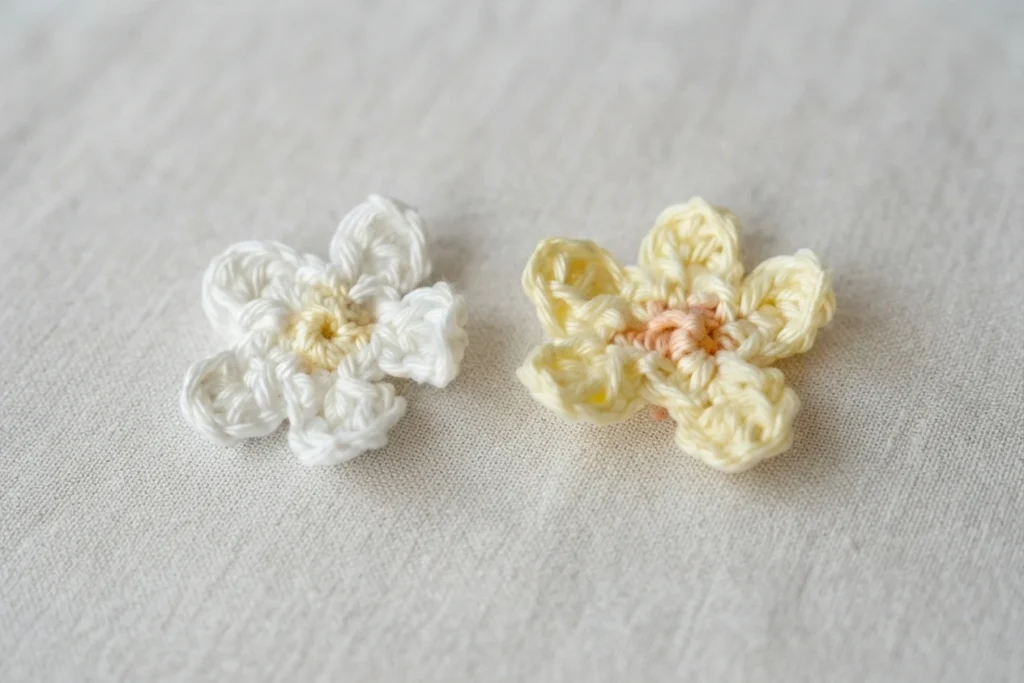 Deux petites fleurs crochetées, l’une blanche à cœur jaune et l’autre crème à cœur pêche, posées sur un tissu clair.