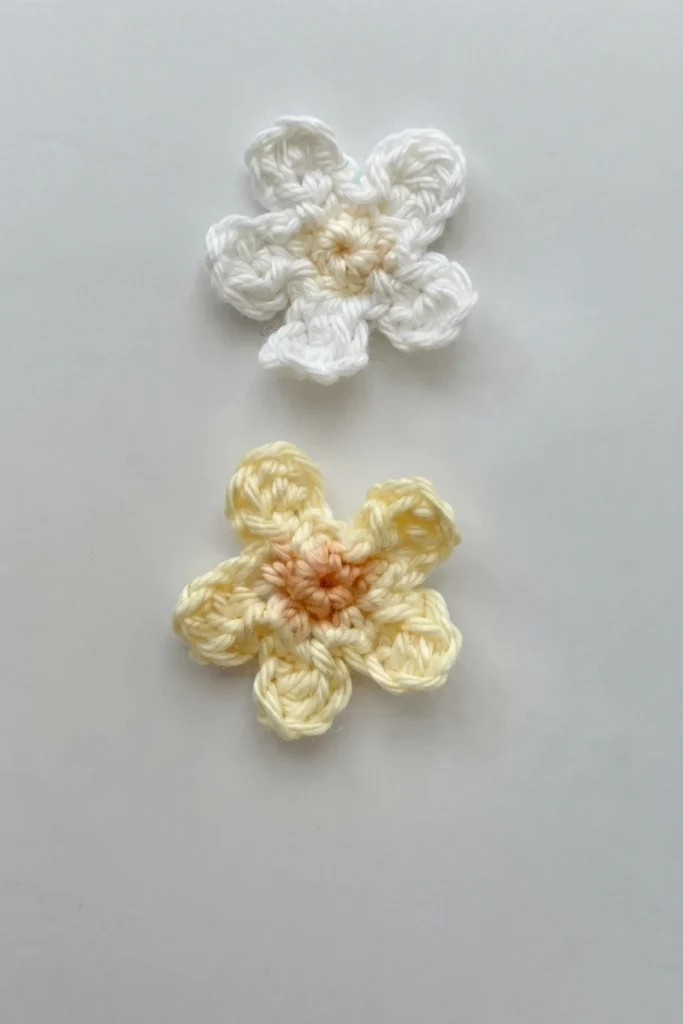 Two crocheted flowers on a light gray surface, one white with a pale yellow center and one pale yellow with a peach center.