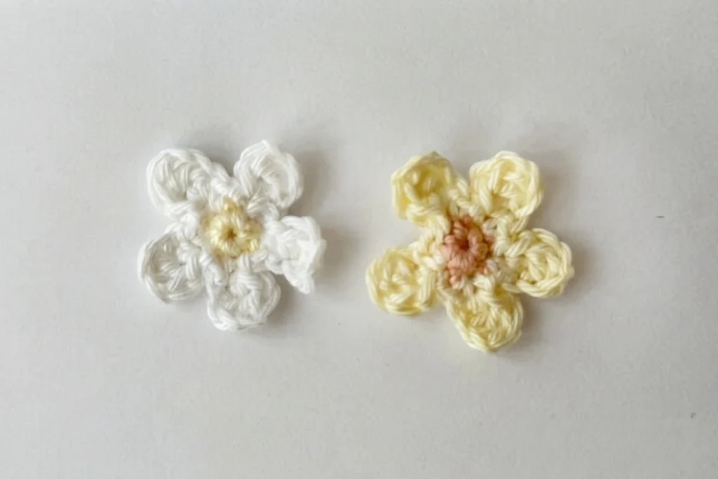 Two small crocheted flowers, one white with a pale yellow center and one pale yellow with a peach center, on a light background.