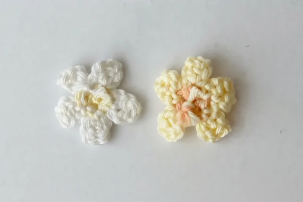 Deux fleurs en crochet, l’une blanche avec un centre jaune pâle, l’autre jaune pâle avec un centre corail, posées sur un fond clair.
