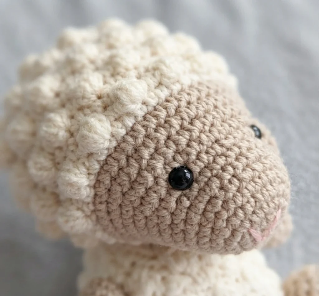 Close-up of a crocheted lamb head with cream bobble-stitch wool, a beige face, black safety eyes and a small embroidered pink mouth.