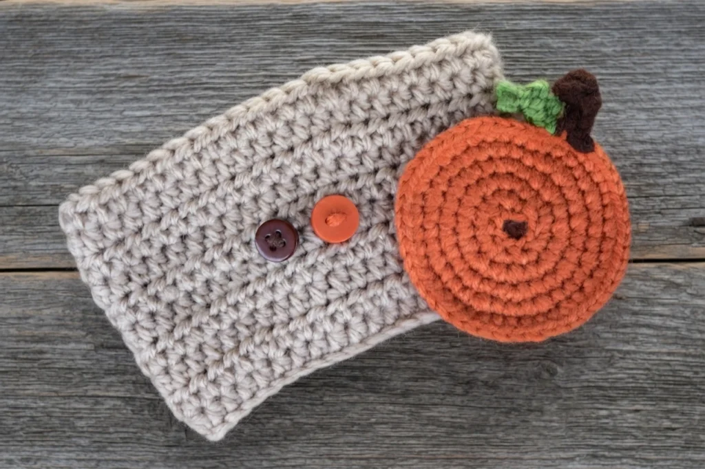 Crocheted cream-colored cup sleeve with two buttons, one brown and one orange, and a round orange crocheted pumpkin appliqué with a green leaf and brown stem, lying on weathered wood.
