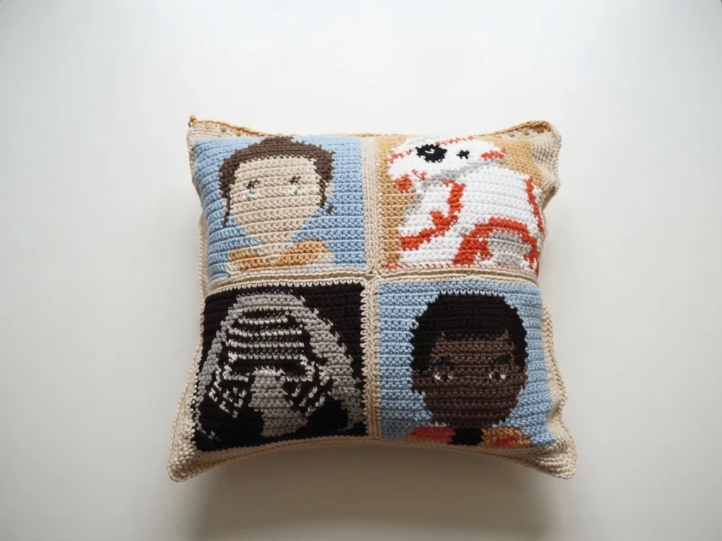 Crocheted Star Wars cushion made of four pixel art squares showing Rey, BB-8, Kylo Ren and Finn, photographed on a plain light background.