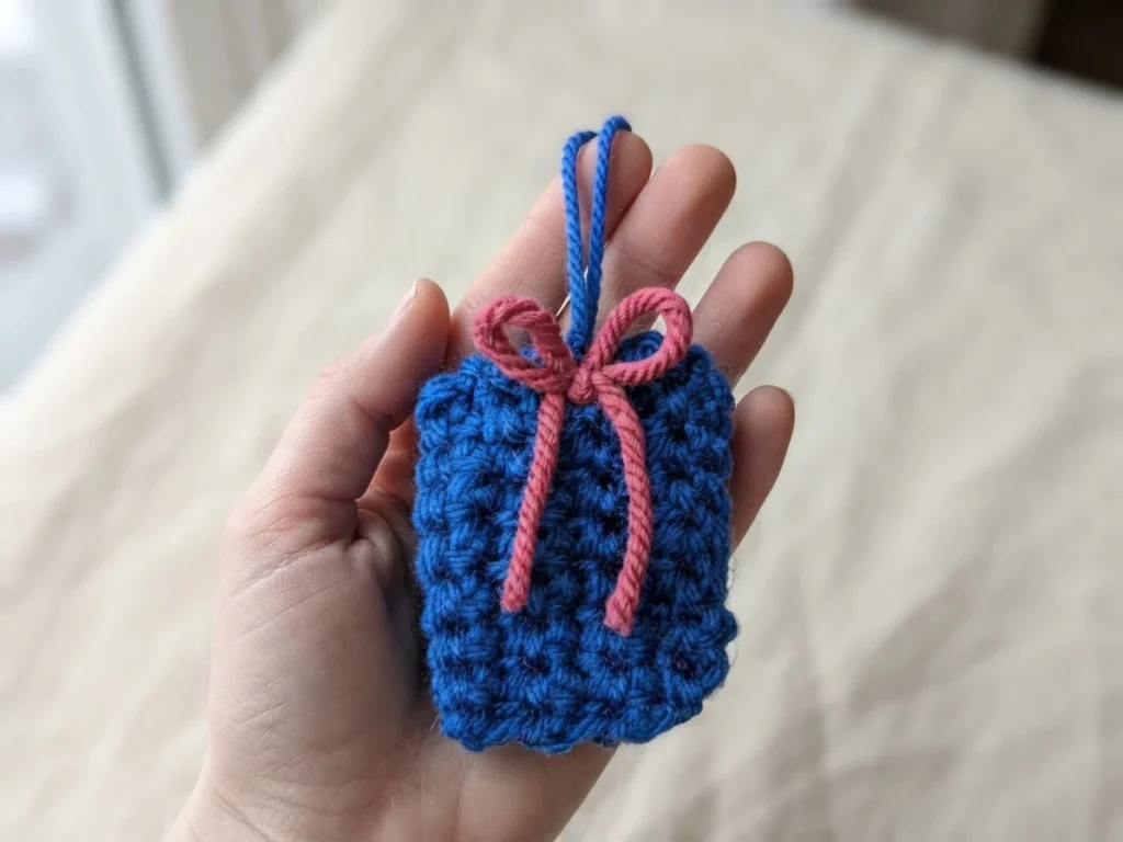 A blue crocheted gift ornament with a pink bow and hanging loop resting in an open hand against a light background.