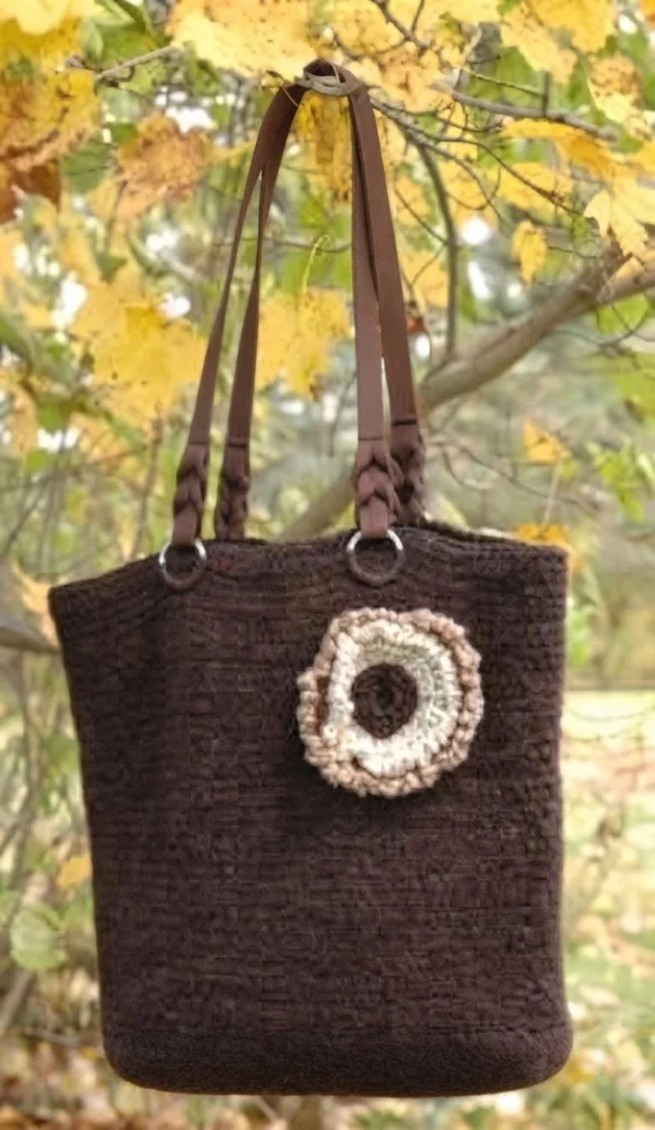 Dark brown crochet tote bag with long straps and a raised cream-and-tan circular motif, photographed among autumn leaves.